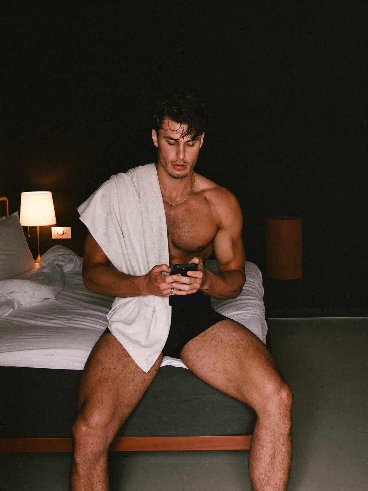 A male creator in black boxers sitting on the edge of a bed with his phone